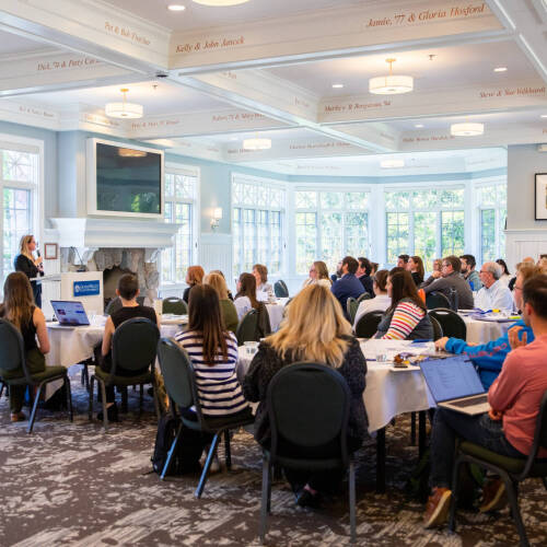 large crowd seated at round tables listens to Stacie Behler at front of room